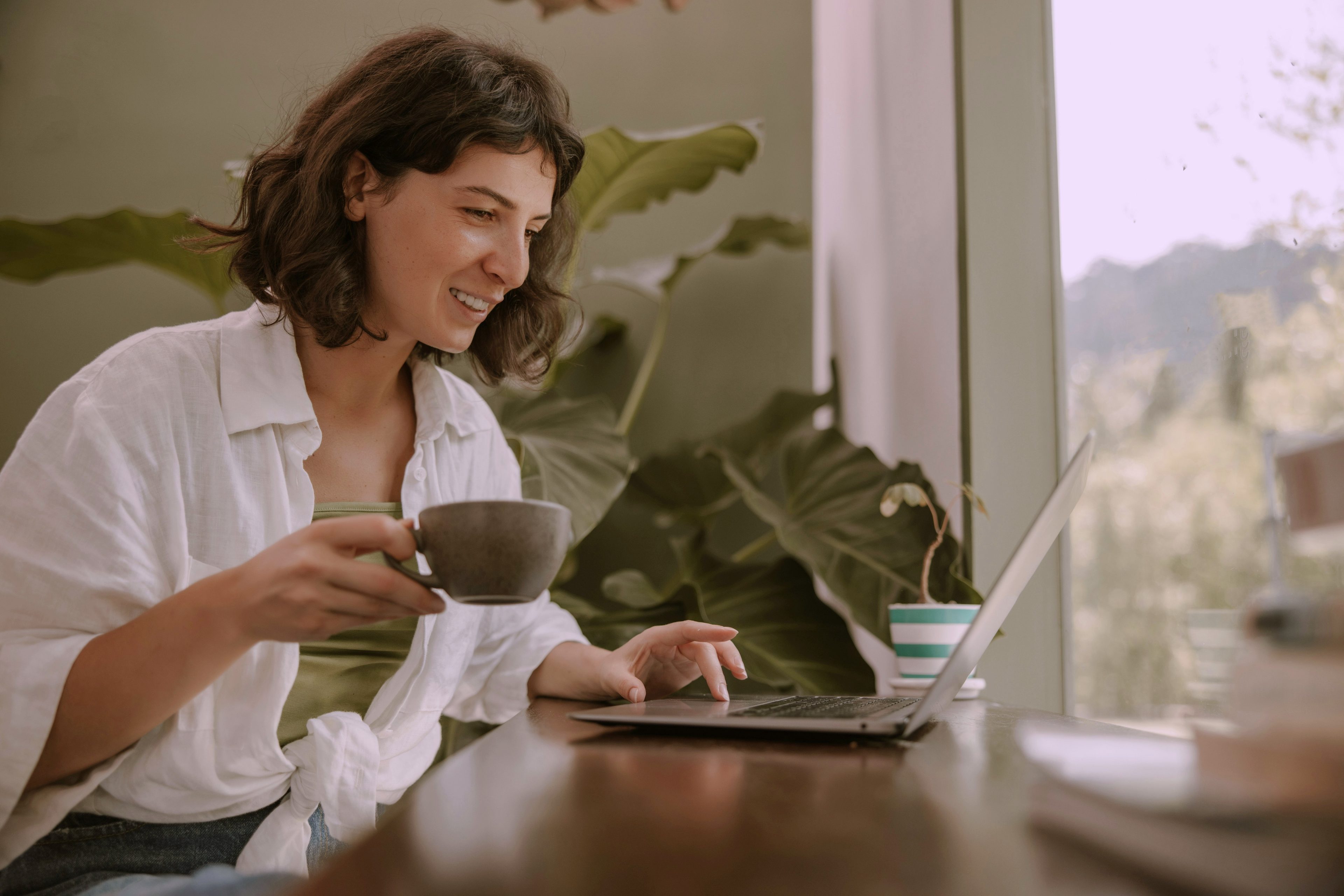 A woman smiling at her laptop with a coffee cup in hand, working at a wooden desk beside a large leafy plant